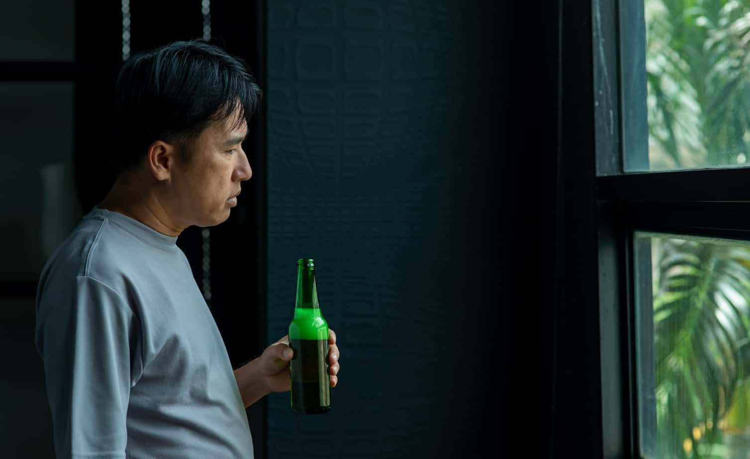 Depressed middle age Asian man standing in the room with drinking beer and think about life problem. Stressed unemployed guy feel upset and tired at home. Mental health and alcohol addiction and economic problem concept.
