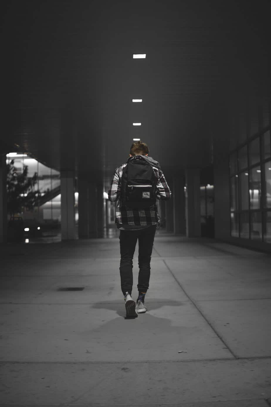 A man walks alone in a dark outdoor walkway from behind, a black backpack on.