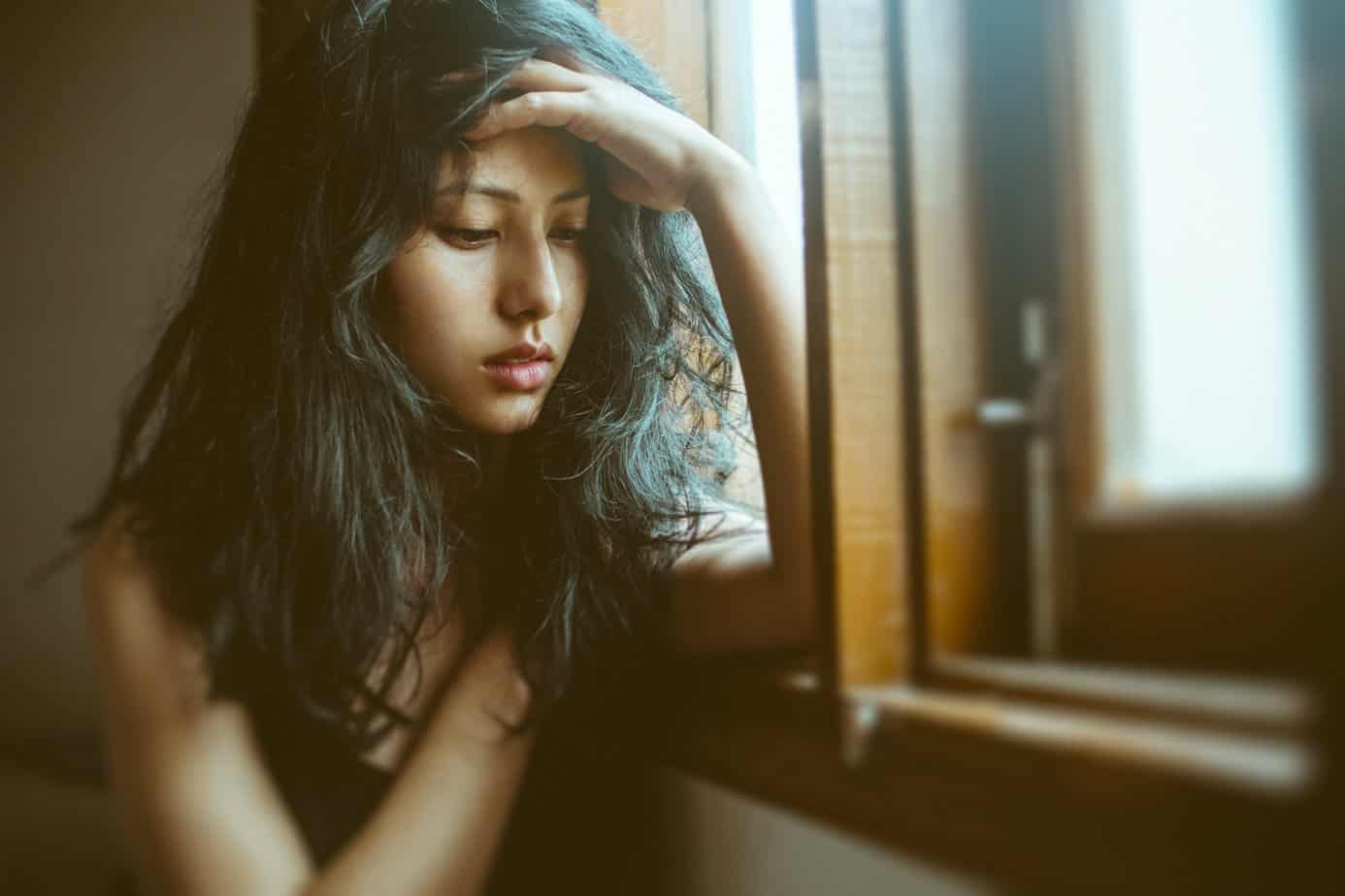 distressed young woman sits by window with head in hands