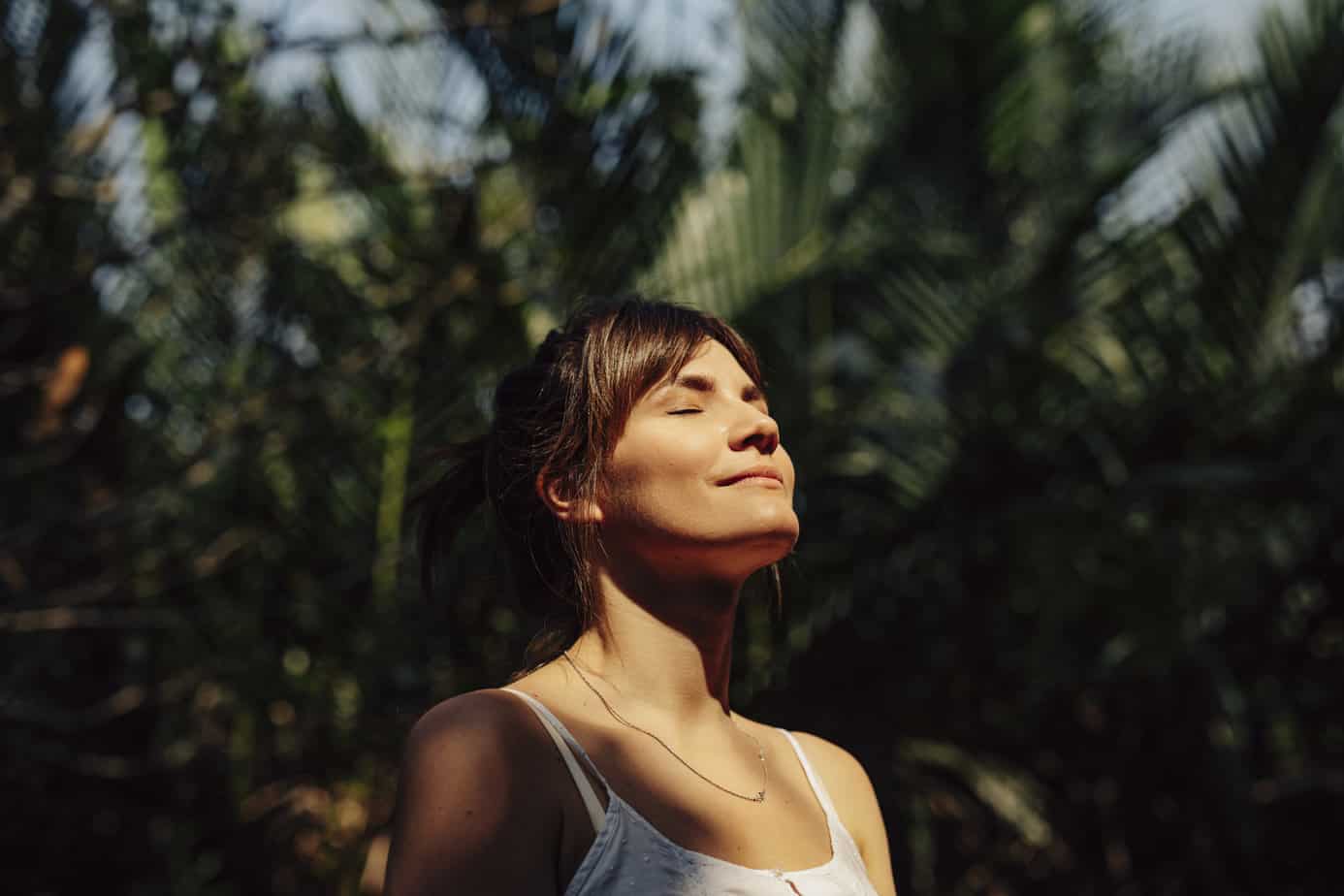 woman with eyes closed standing in sunlit forest and practicing meditation