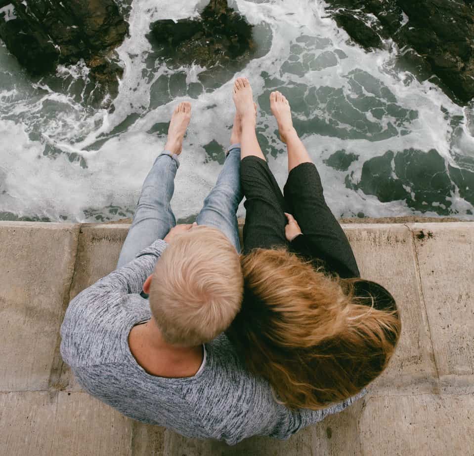 Couple in sweatpants barefoot sitting on pier above ocean