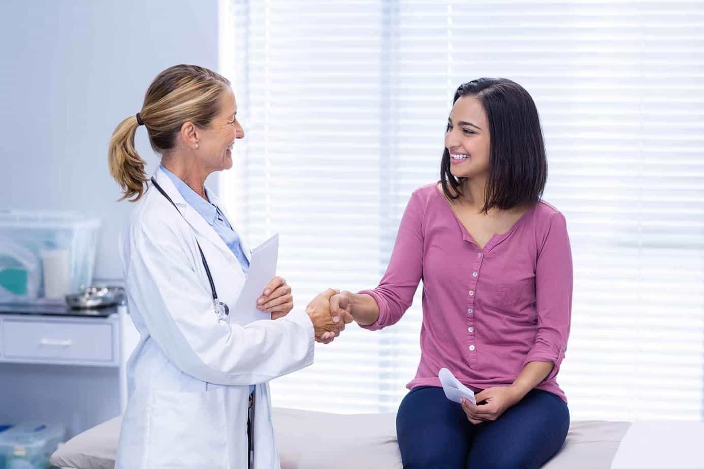 Female client sitting on medical examination table shaking hands with a doctor
