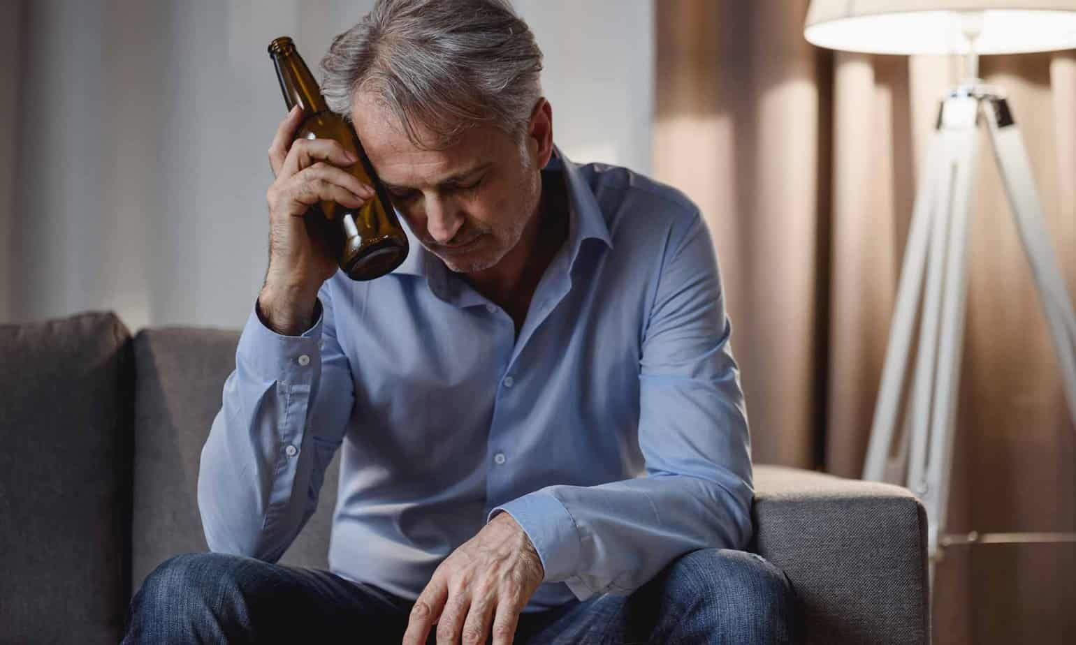 Older gray haired man in blue jeans and blue shirt holding beer bottle to head looking sad on living room couch