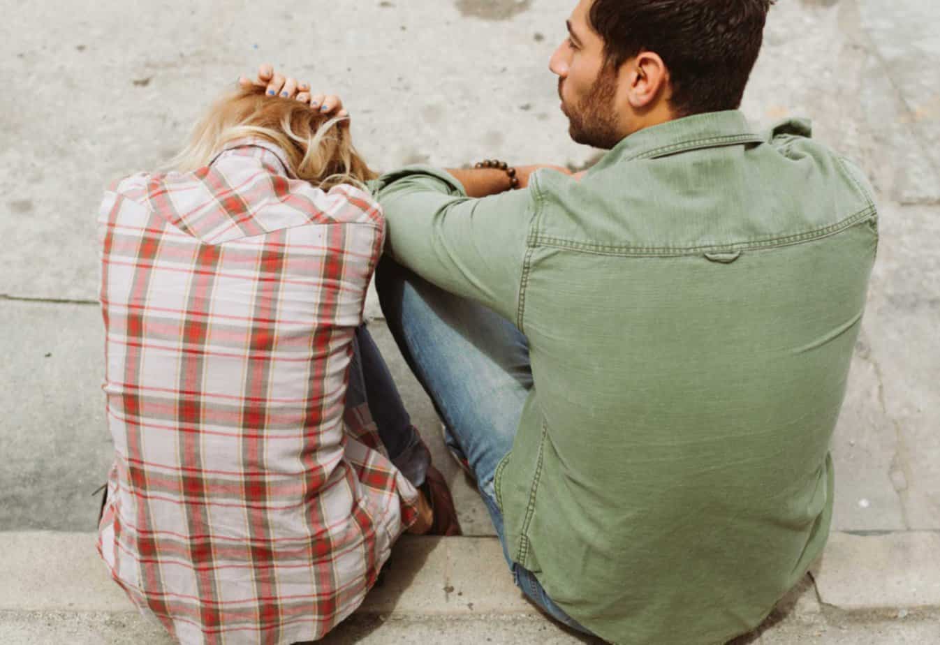 frustrated woman in flannel with head in arms on outside stoop next to man in green jacket