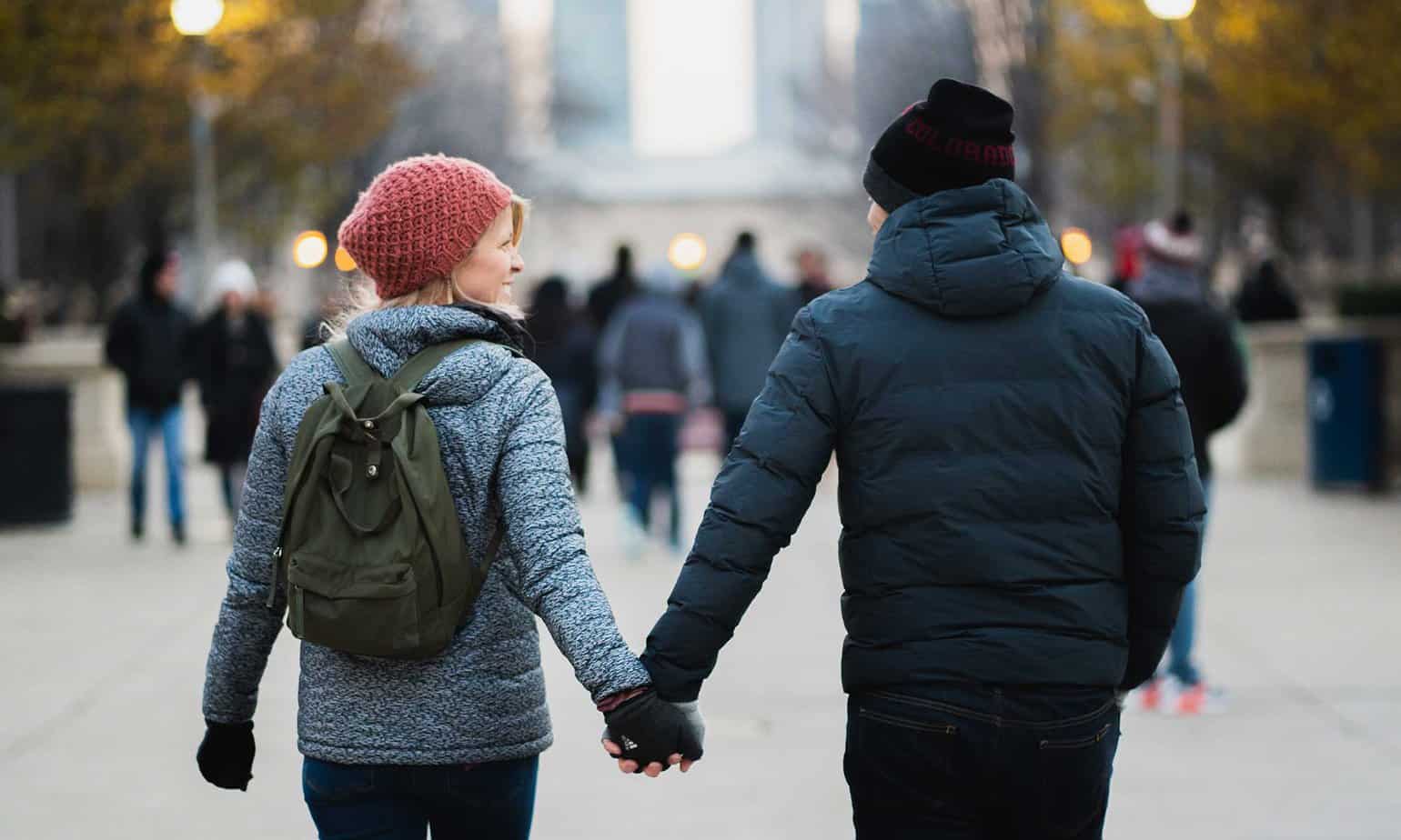 man and smiling woman in blue jackets and hats holding hands walking in a city on a cold day