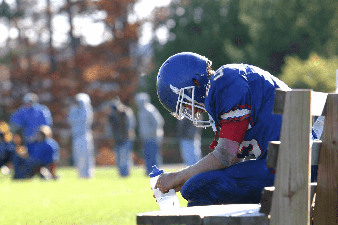 football player on bench holding a water bottle looking sad while team plays on field