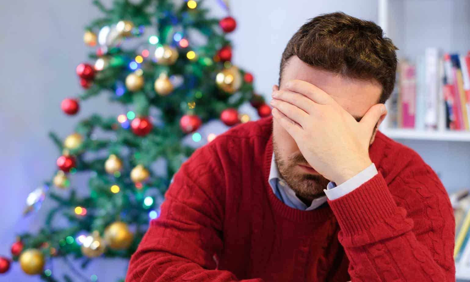 Man stressing over the holidays with hand on head and Christmas tree in the background