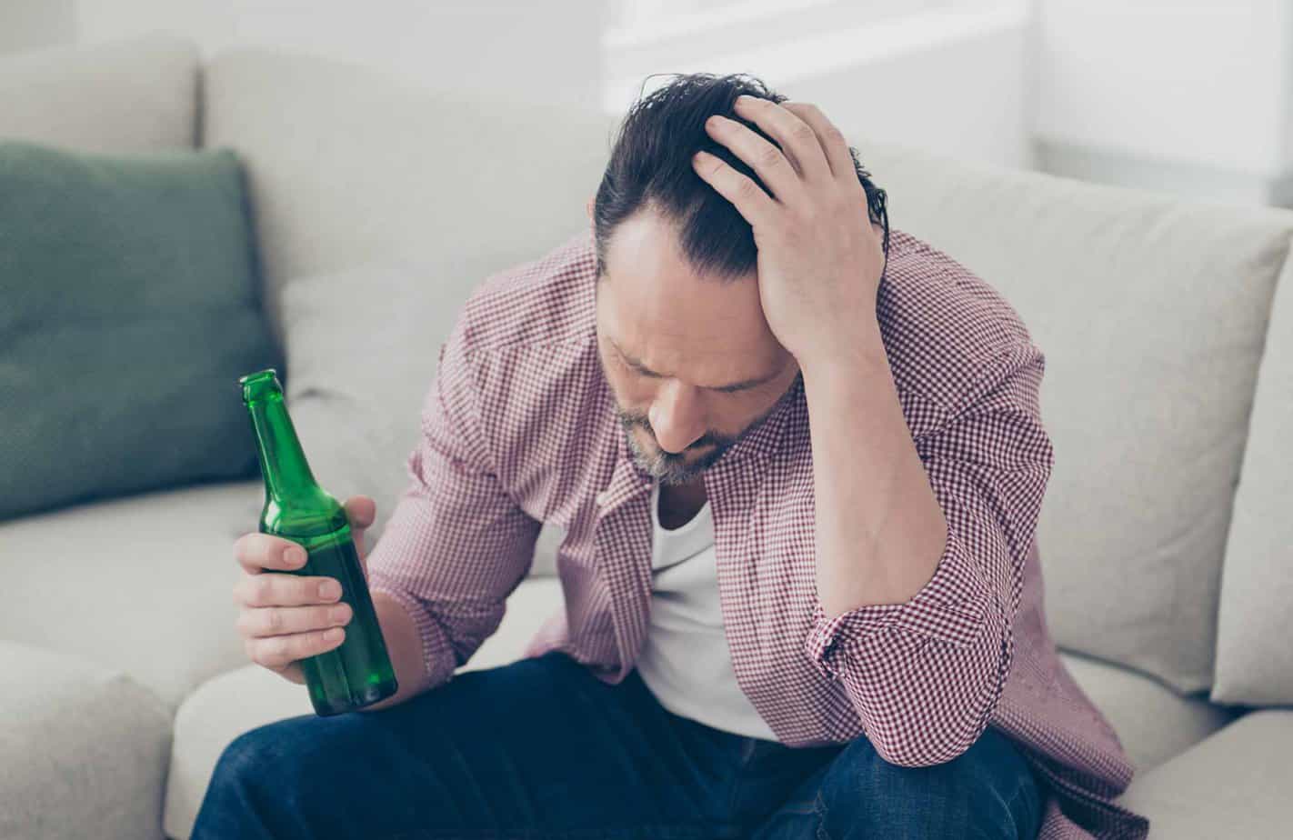 man holding head in hand with beer bottle in other sitting on couch
