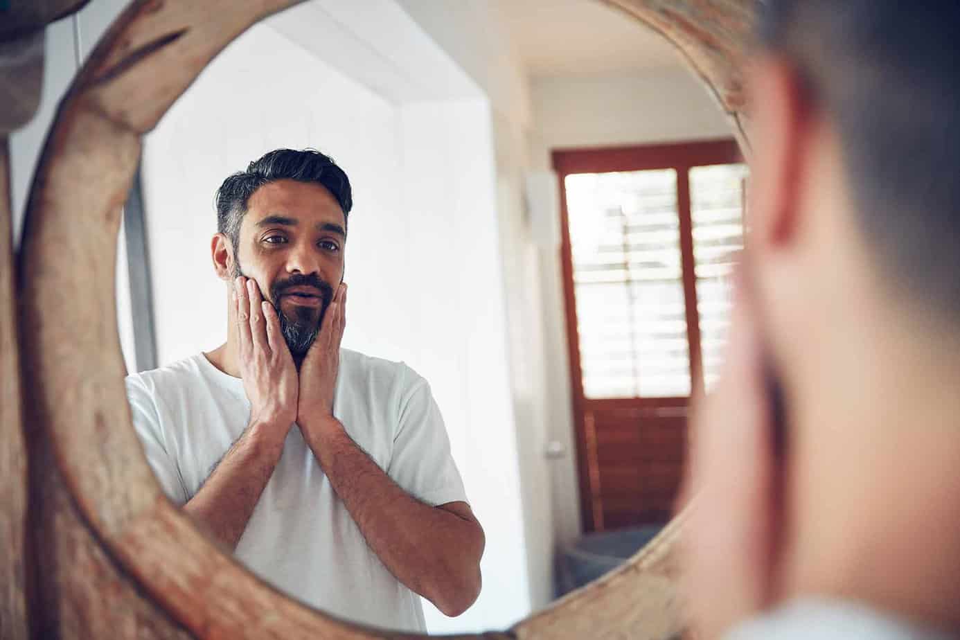 man with hands on face looking into wooden mirror