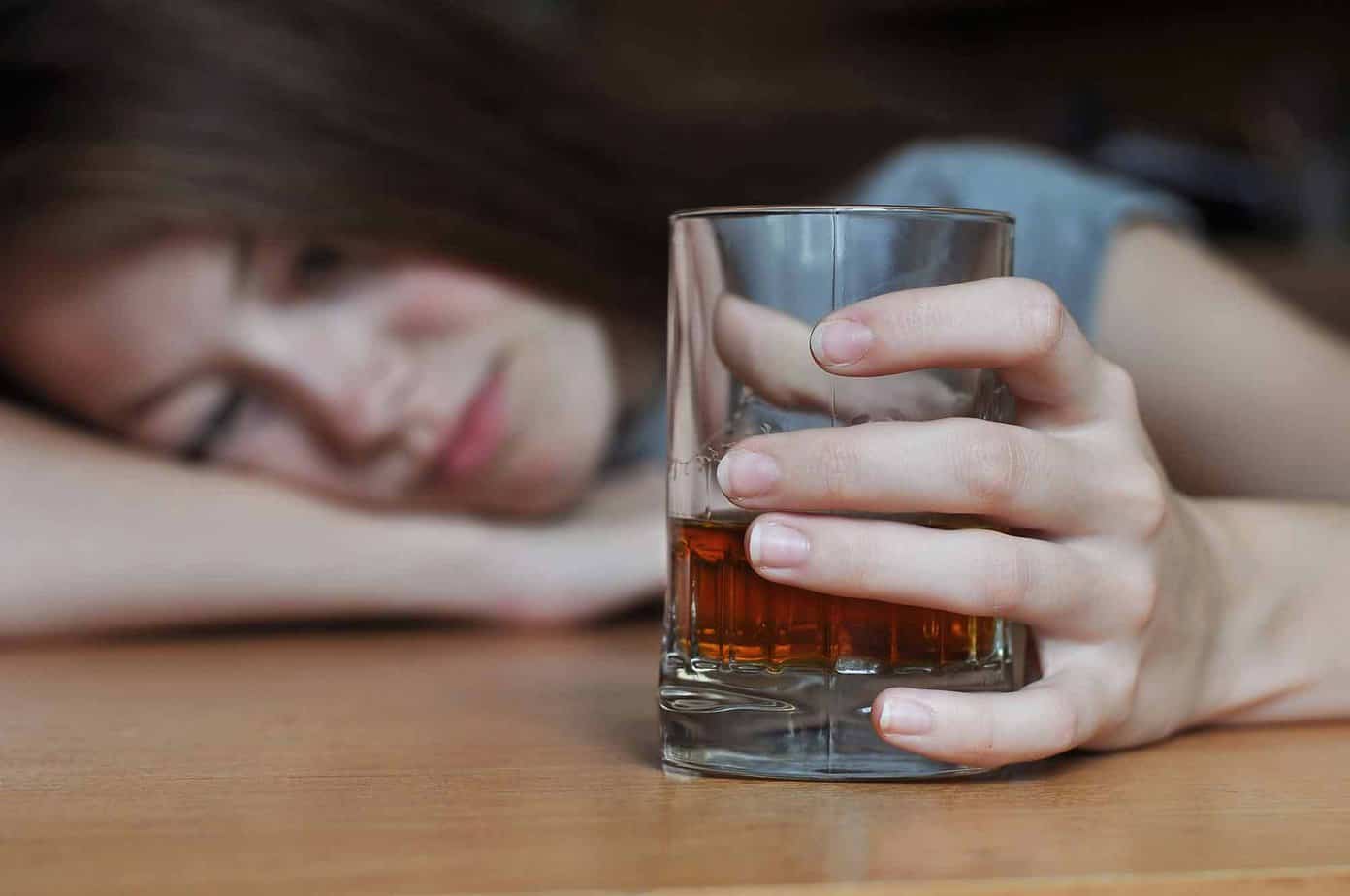 depressed woman lies on table clutching a glass containing liquor