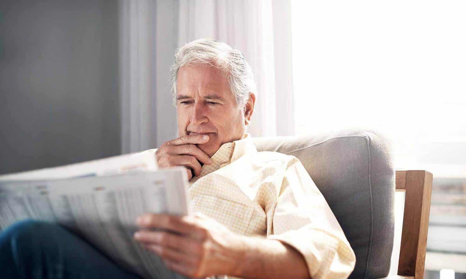 An older man in a yellow dress shirt, reading a newspaper - Stock