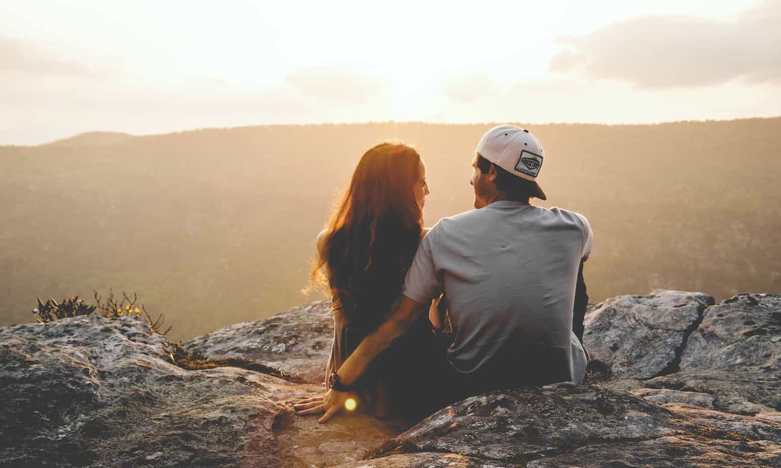 man and woman sitting romantically on cliffside watching sunset