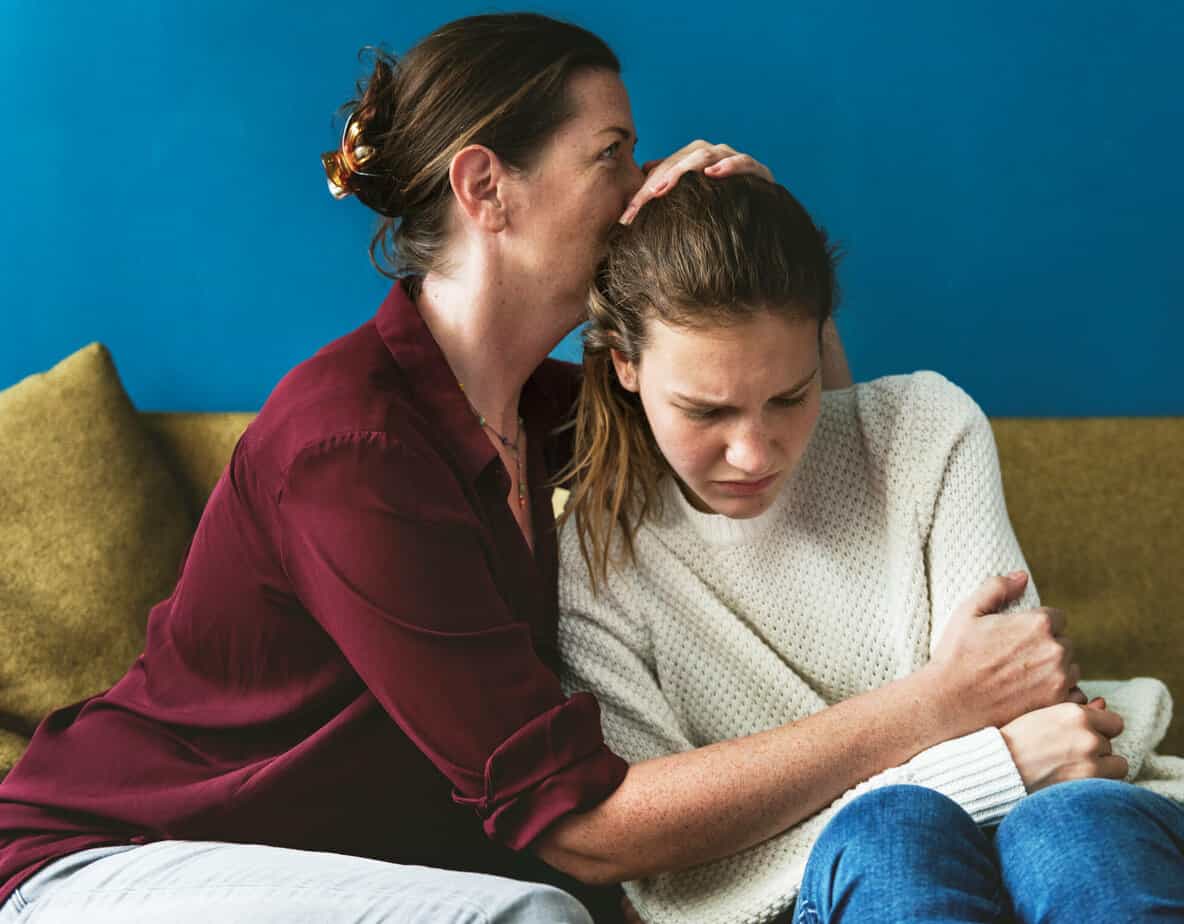 mother comforting crying daughter with arm around her on couch