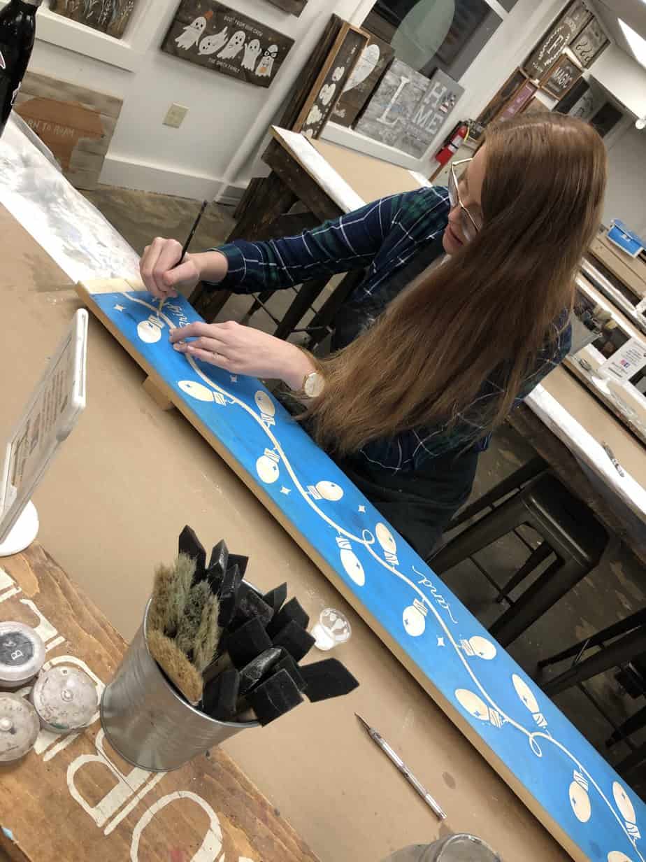 Woman painting wooden board with christmas holiday lights at Mountainside Treatment Center Extended Care Board & Brush Event October 2022