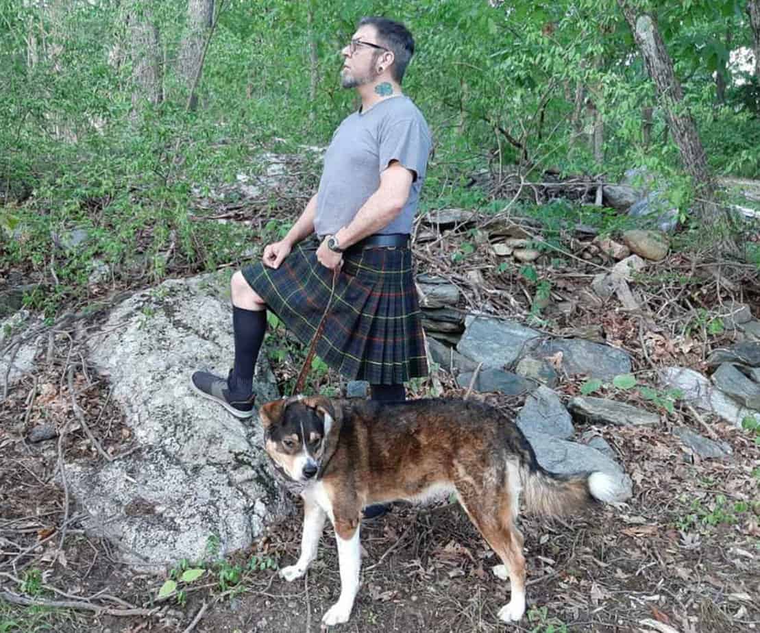 man in kilt stands on top of rock with dog