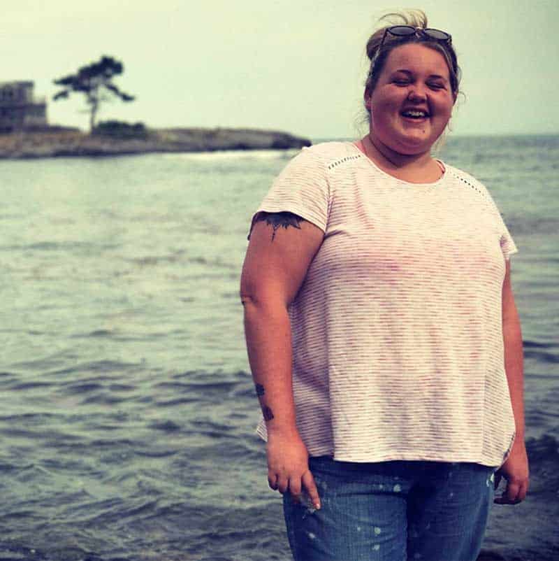 Happy woman in pink shirt and blue jeans stands next to body of water