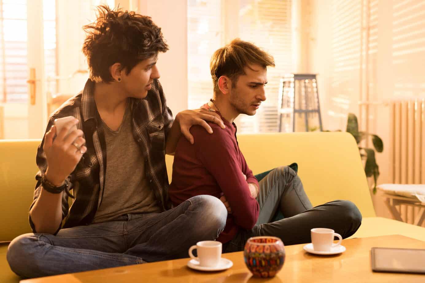 gay couple seated on couch, one partner comforts other partner who looks upset