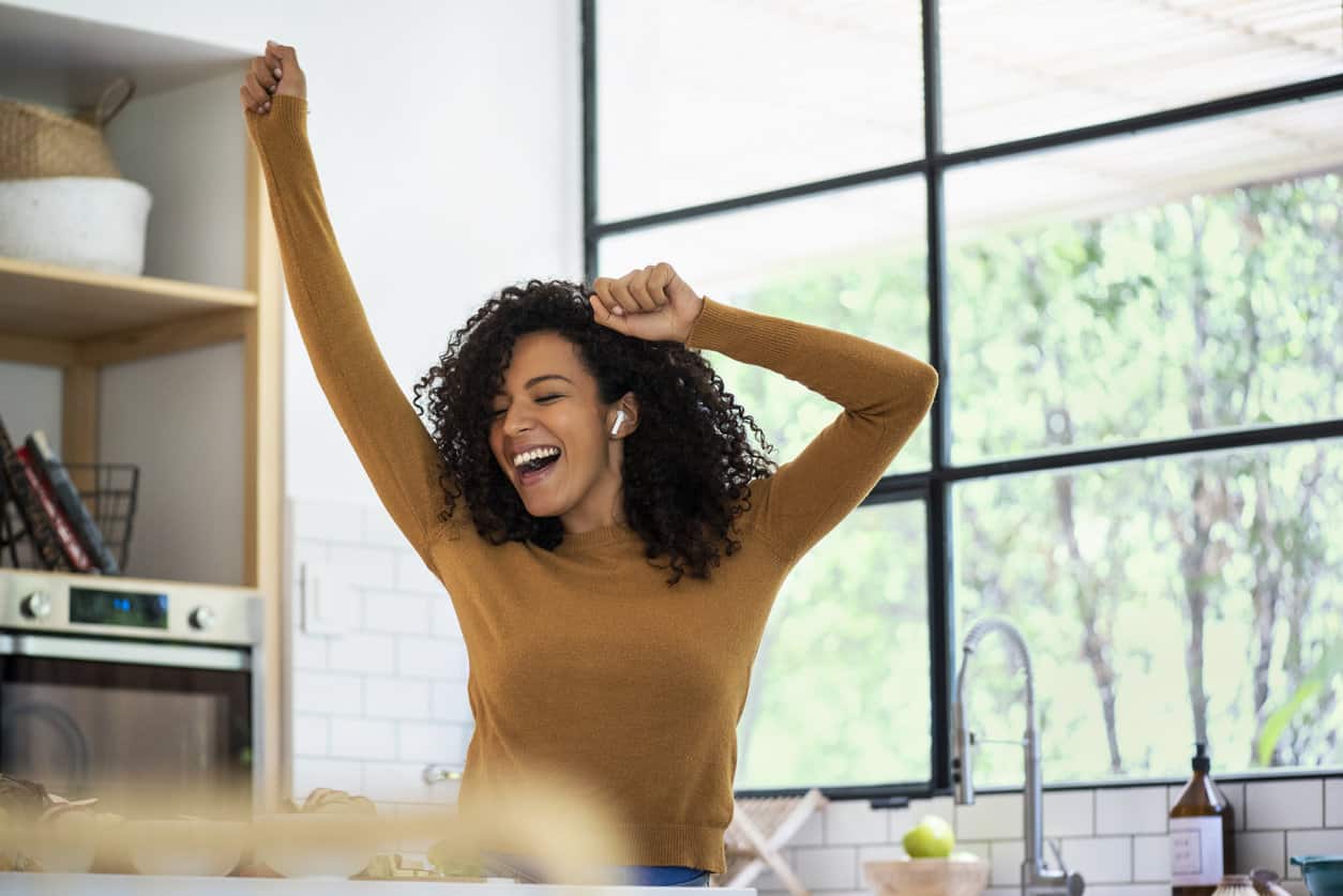 Happy woman with eyes closed listening music on earbuds and dancing in kitchen