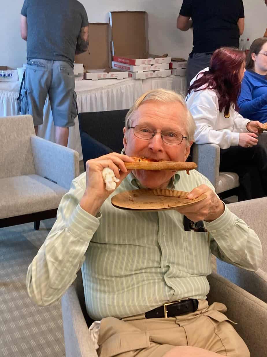 Older man sitting down smiling while eating a slice of pizza at Alumni 2023 Pizza Social at Canaan